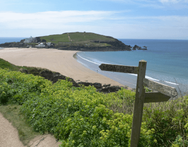 A fingerpost on the South West Coast Path points across a beach exposed by low tide to Burgh Island in the distance.